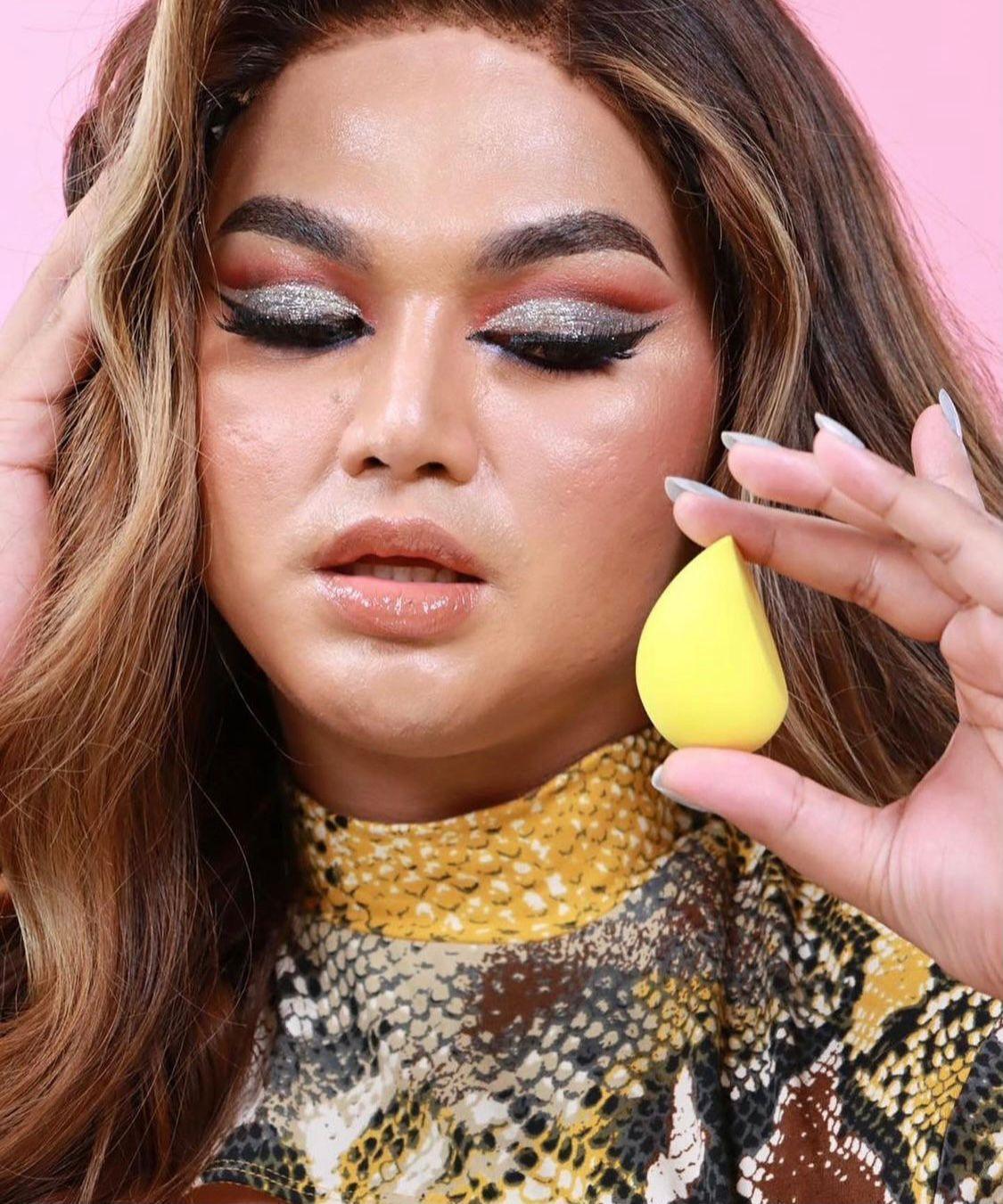 Person with glamorous makeup holding yellow teardrop makeup sponge, snake print top
