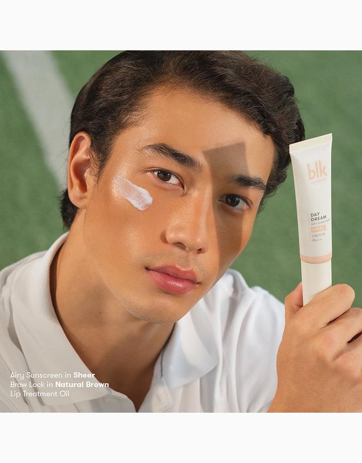 Man applying sheer sunscreen to face, holding blk Daydream sunscreen tube on green background