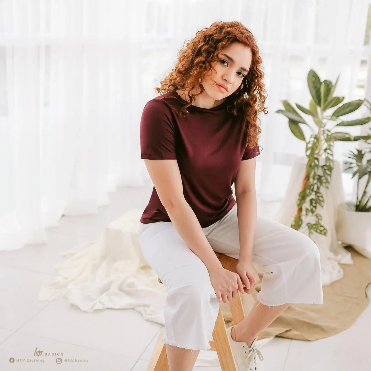 Woman in maroon slim fit t-shirt and white cropped pants sitting on stool in bright room