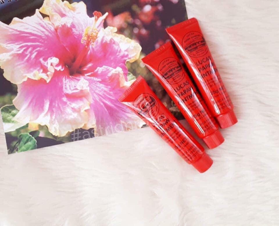 Three red Lucas Papaw Ointment tubes on a soft surface beside a pink hibiscus flower photo.