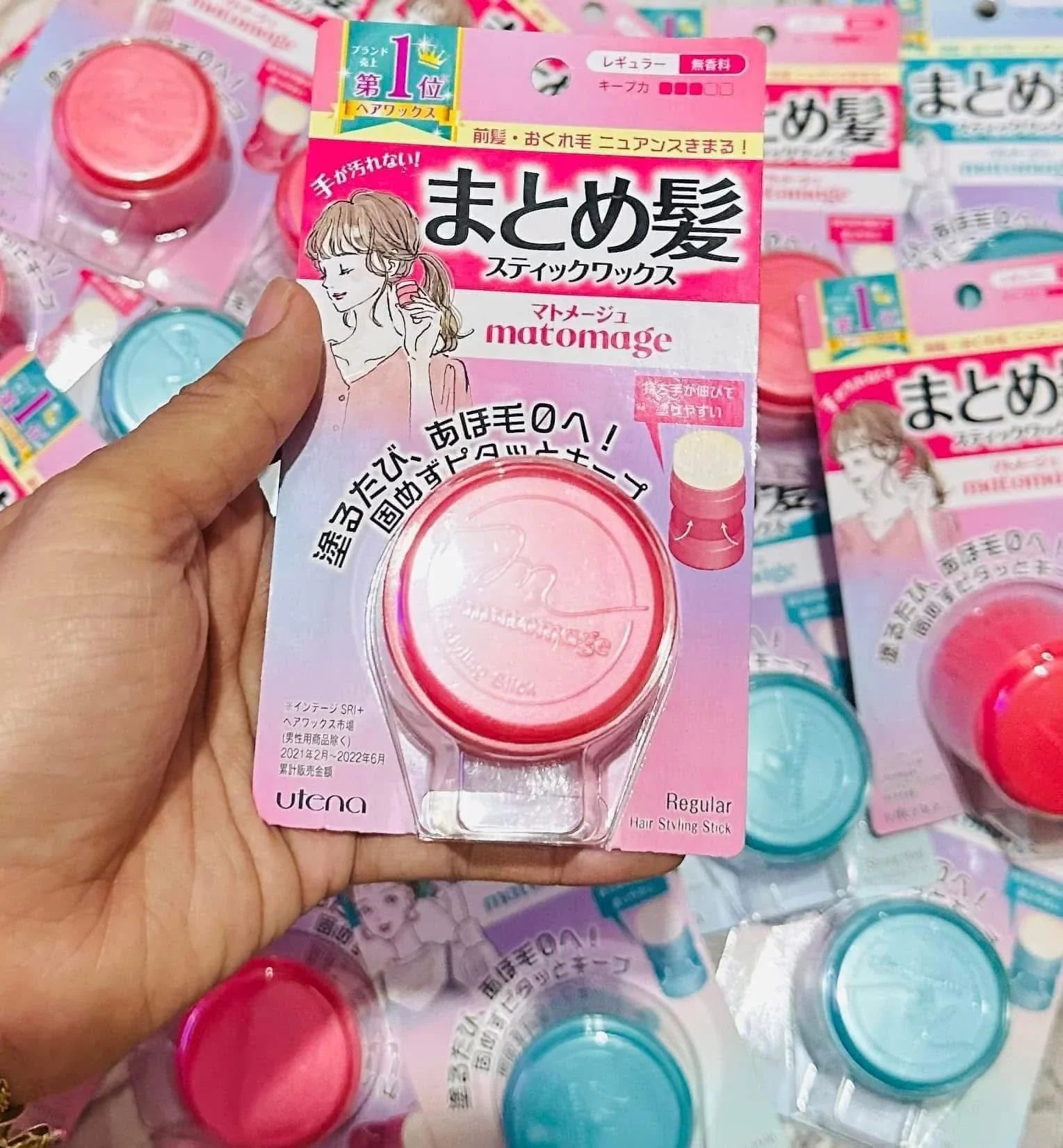 Hand holding pink Matomage hair styling stick in Japanese packaging, surrounded by similar products.