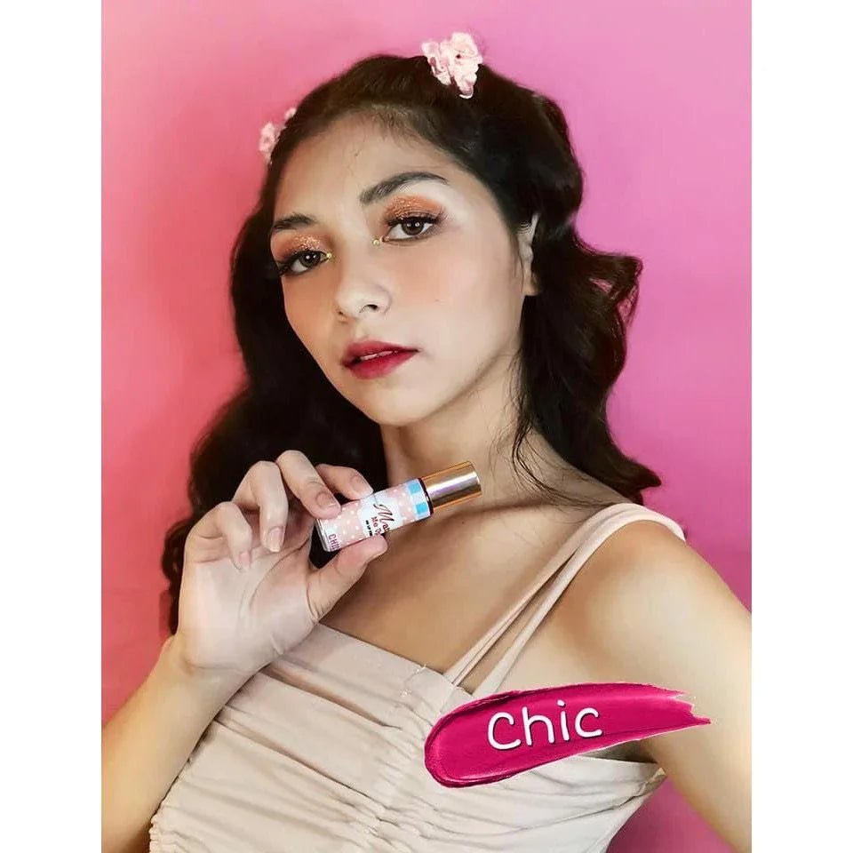 Woman with flower hair clips holding matte liquid lipstick, pink background, 'Chic' shade.